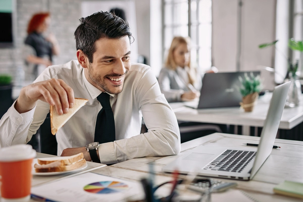 A young man sitting on his desk in office and eating snacks.